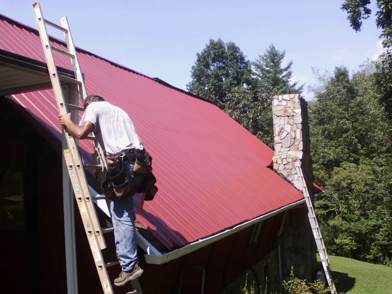 Roofer on a ladder beside a red metal roof and stone chimney — Soffit & Fascia Repair by Roofing Brothers in Tendoy